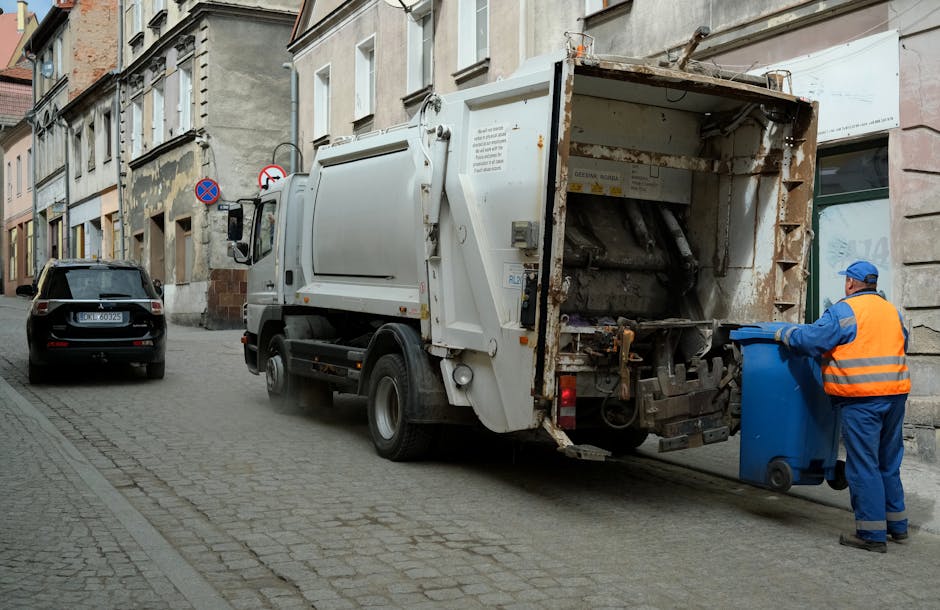 A waste collection truck, painted white with visible signs of rust and dirt, is parked on a narrow cobblestone street in an urban area lined with old multi-storey buildings with beige and gray facades. The truck's rear chute is open, revealing mechanical components used for compacting and lifting rubbish. A worker dressed in a blue uniform with an orange high-visibility vest and matching cap is positioned to the right, actively pushing a blue wheeled bin toward the truck’s intake opening. The environment appears to be a typical city street with minimal traffic, as evidenced by a small black car parked further down the street and traffic signs, including a no-parking sign mounted on a pole attached to one of the buildings. The lighting suggests an overcast day, providing diffuse natural illumination. This scene illustrates a routine private waste collection process, reflecting alternative rubbish handling methods often used in conjunction with or separate from local authority services, and is typical for a professional rubbish removal service such as Rubbish Removal Hammersmith.