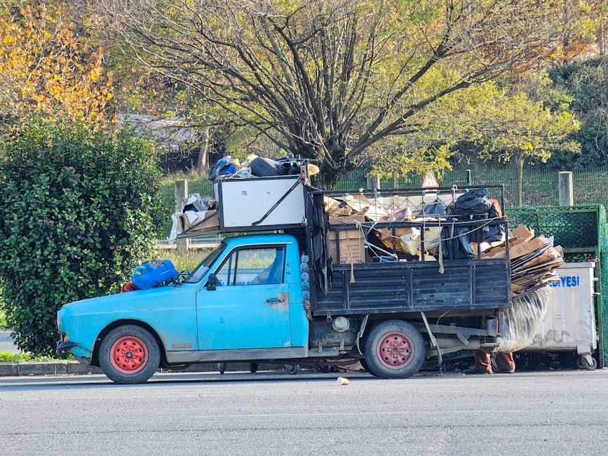 A small, light blue, two-door utility truck is parked on the side of a paved road in an outdoor environment. The vehicle's paint appears worn and slightly faded, with visible rust and dirt on the front fender and sides. The truck features red painted steel wheels and black side mirrors. The truck bed is loaded with an overflowing collection of assorted waste materials, including large cardboard boxes, black plastic garbage bags, and miscellaneous scrap items, which are secured with ropes and straps. Some waste is piled high, extending beyond the sides of the truck bed. Behind the vehicle, there is a large deciduous tree with sparse green and yellow leaves, suggesting an autumn setting. To the left of the truck, a dense green bush is partially visible, and further in the background, a fence with vertical metal posts and a leafy hedge can be seen, enclosing a garden or park area. The scene is illuminated by natural daylight, casting soft shadows, and the overall context suggests an on-site rubbish collection or alternative waste handling service provided by Rubbish Removal Hammersmith.