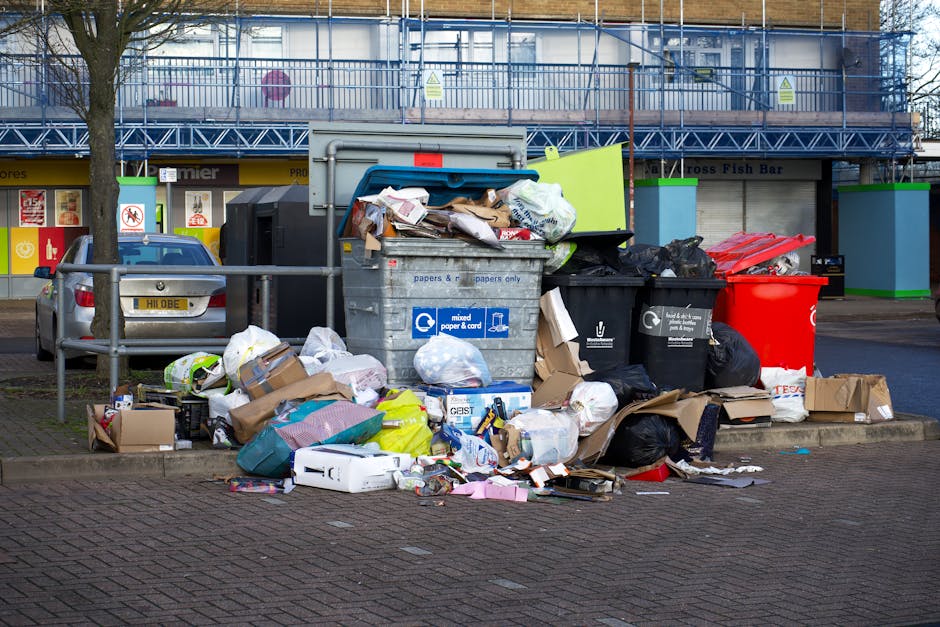 A waste collection truck, painted white with visible signs of rust and dirt, is parked on a narrow cobblestone street in an urban area lined with old multi-storey buildings with beige and gray facades. The truck's rear chute is open, revealing mechanical components used for compacting and lifting rubbish. A worker dressed in a blue uniform with an orange high-visibility vest and matching cap is positioned to the right, actively pushing a blue wheeled bin toward the truck’s intake opening. The environment appears to be a typical city street with minimal traffic, as evidenced by a small black car parked further down the street and traffic signs, including a no-parking sign mounted on a pole attached to one of the buildings. The lighting suggests an overcast day, providing diffuse natural illumination. This scene illustrates a routine private waste collection process, reflecting alternative rubbish handling methods often used in conjunction with or separate from local authority services, and is typical for a professional rubbish removal service such as Rubbish Removal Hammersmith.