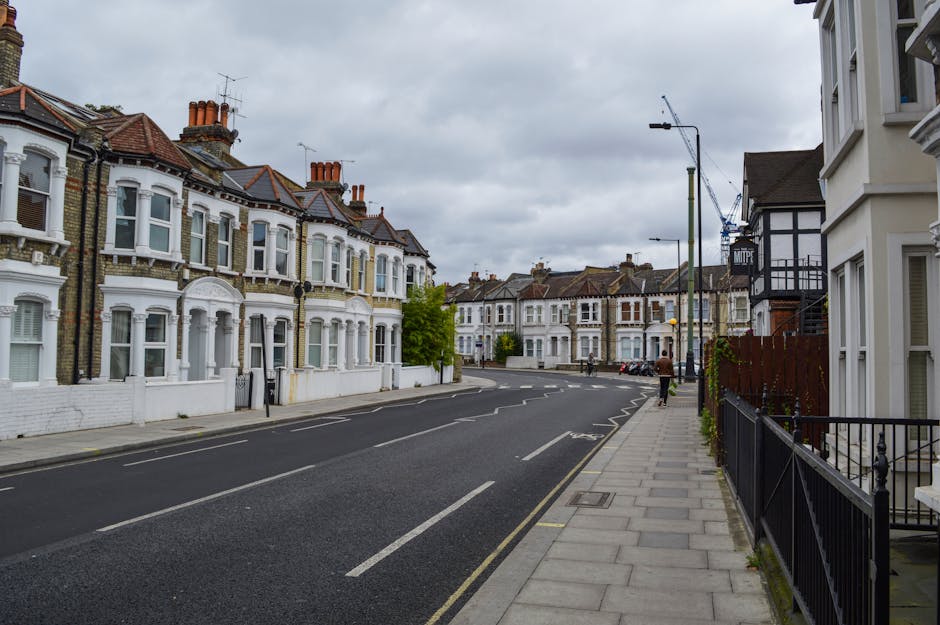 A quiet residential street featuring a row of Victorian-style terraced houses with brick facades and white-painted bay windows, situated on the left side of the image. The houses have red-tiled pitched roofs adorned with multiple chimneys and small decorative architectural elements above the windows. On the right side, part of a white building with black timber framing is visible, alongside a black metal fence bordering a small garden area. The paved sidewalk runs parallel to the street, with a few pedestrians walking along it. Streetlights and a construction crane are seen in the background, indicating ongoing development. The overcast sky casts diffuse light across the scene, with a few parked cars visible in the distance. The overall scene captures a typical late 20th-century residential area, relevant to waste management and rubbish removal services that focus on local household waste collection or private clearance in suburban neighborhoods.