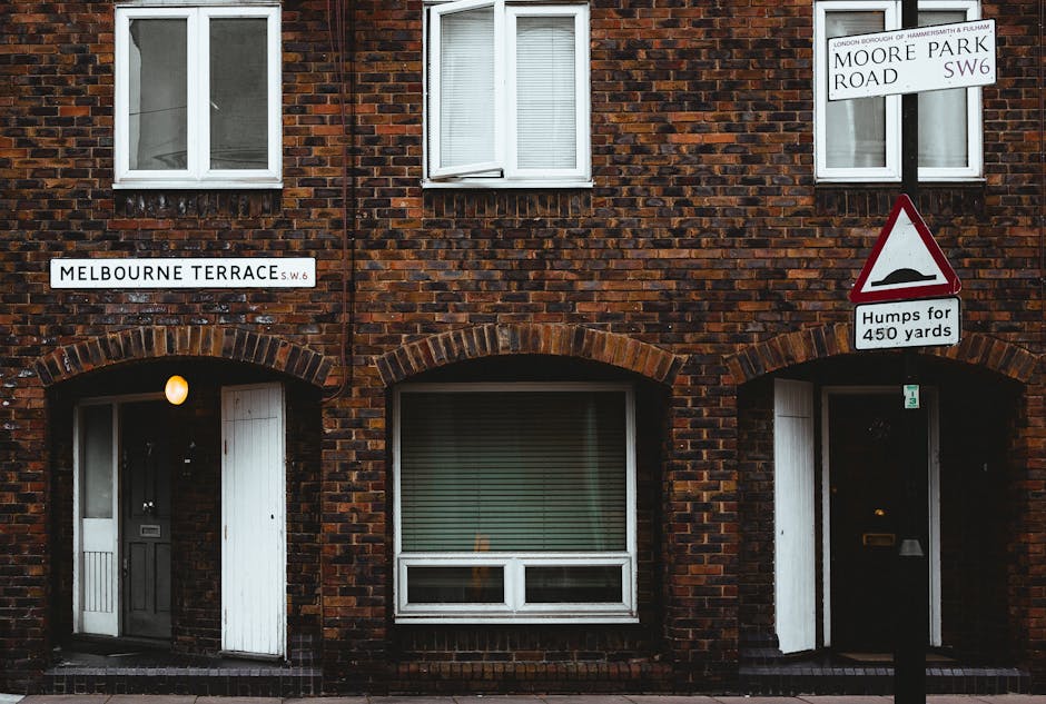 A quiet residential street featuring a row of Victorian-style terraced houses with brick facades and white-painted bay windows, situated on the left side of the image. The houses have red-tiled pitched roofs adorned with multiple chimneys and small decorative architectural elements above the windows. On the right side, part of a white building with black timber framing is visible, alongside a black metal fence bordering a small garden area. The paved sidewalk runs parallel to the street, with a few pedestrians walking along it. Streetlights and a construction crane are seen in the background, indicating ongoing development. The overcast sky casts diffuse light across the scene, with a few parked cars visible in the distance. The overall scene captures a typical late 20th-century residential area, relevant to waste management and rubbish removal services that focus on local household waste collection or private clearance in suburban neighborhoods.