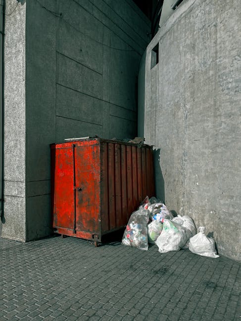 A large, weathered red metal rubbish skip with visible rust and peeling paint, positioned against a dark grey, smooth-clad wall on the left and a textured, light grey stone wall on the right, located outdoors on a paved grey surface. The skip is partially filled with waste, and several white and transparent plastic bags containing mixed rubbish are piled on the ground beside it, leaning against the side of the skip. The environment appears to be an alleyway or service area, with minimal natural light filtering in from above, creating subtle shadows around the containers and bags. This scene exemplifies a typical private waste collection scenario that may be handled by companies such as Rubbish Removal Hammersmith, often used as an alternative to council rubbish collection services, particularly for larger quantities of waste generated from commercial or renovation activities.