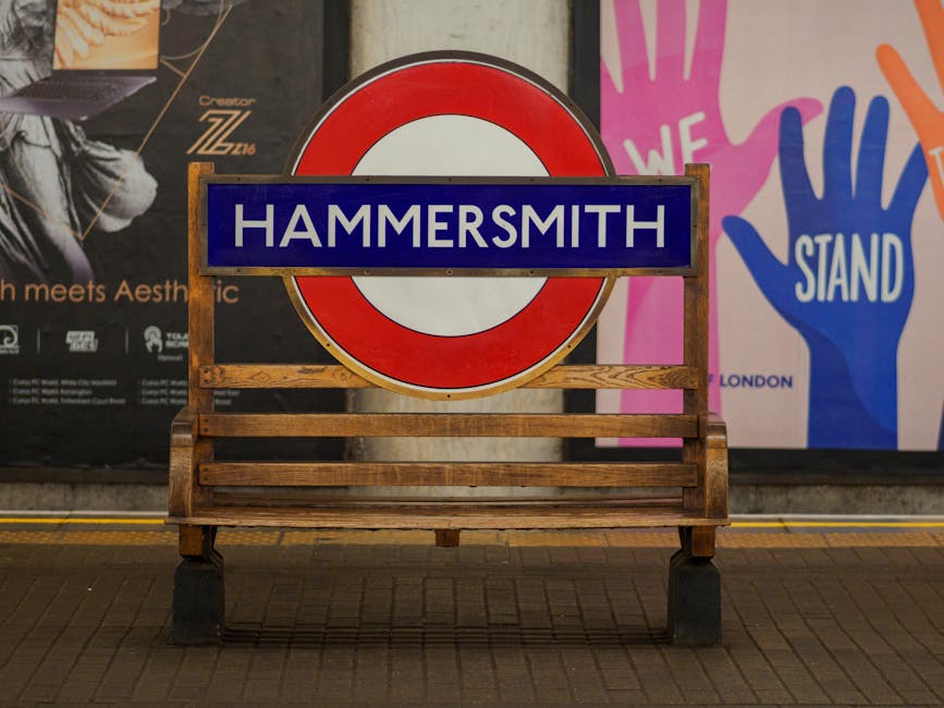 A wooden bench with horizontal slats and sturdy black metal legs is positioned on a tiled surface in front of a large circular London Underground sign with a red and white ring and a blue rectangular panel reading 'HAMMERSMITH'. Behind the sign, there are colorful posters including one with prominent hand graphics and the word 'STAND' in white letters on a blue hand outline, and another partially visible featuring black, gold, and white elements. The setting appears to be an indoor or covered station area, illuminated with artificial lighting, and the background showcases posters and advertisements on a wall. The scene suggests a location in Hammersmith related to public transit, which aligns with the context of alternative waste disposal or rubbish collection services provided by Rubbish Removal Hammersmith in the area.