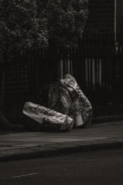 Two large black plastic sacks filled with household waste, positioned on a narrow sidewalk near a black metal fence with vertical bars and a lattice panel behind it. The sacks are crumpled and appear to contain mixed rubbish, with some white and dark contents visible through the thin plastic. They are leaning slightly to the right, resting against each other, and are partially shaded by nearby trees with dense foliage in the background. The setting is an outdoor urban area, with a curb separating the sidewalk from a paved road in the foreground. The overall scene is dimly lit, suggesting overcast weather or early evening, and reflects a typical private waste disposal solution that might be arranged through a rubbish removal service such as Rubbish Removal Hammersmith, independent of local authority collection. The environment is clean aside from the waste sacks, emphasizing the importance of proper rubbish management in residential or commercial properties to prevent clutter and promote safety.