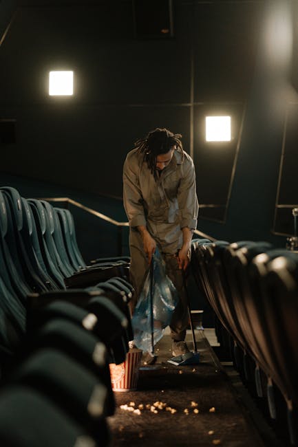 A person with dreadlocks wearing a grey jumpsuit is cleaning up trash and debris from the floor of an auditorium or theatre seating area. They are collecting waste from a blue plastic bag while standing between dark green upholstered seats arranged in rows on either side. The environment features a dark wall with two bright square lights casting illumination, and a metal railing running along the side of the seating area. The floor appears to be dark and slightly textured, with some scattered litter visible on the ground. The scene suggests a professional cleaning or waste removal activity focused on maintaining the cleanliness of a venue, which aligns with the services provided by Rubbish Removal Hammersmith in alternative waste handling or on-site clearance for commercial or event spaces.