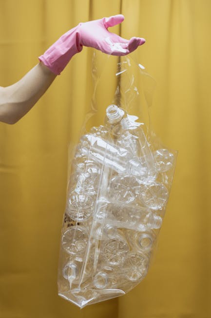 A person wearing a light pink rubber glove is holding a large, clear plastic bag filled with empty transparent plastic bottles, glass bottles, and plastic packaging materials. The background features a plain yellow curtain or wall, providing a neutral backdrop for the image. The person appears to be engaged in a recycling or waste collection activity related to rubbish removal services. The plastic bottles and packaging are loosely contained within the bag, which is being held upright, suggesting it is prepared for disposal or transport. The scene emphasizes the process of sorting and handling recyclable materials, consistent with the context of independent waste clearance or private disposal services provided by Rubbish Removal Hammersmith. The overall lighting is even and bright, highlighting the clarity of the plastic materials and the texture of the gloves, with minimal shadows present.