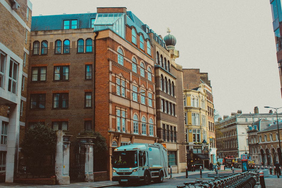 A narrow city street lined with a variety of historic and modern multi-story buildings showcases a mix of brick, stone, and plaster facades in shades of red, tan, and cream. The building in the center is a prominent red brick structure with arched windows on each floor, topped with a decorative cornice and an ornate tower feature with a rounded dome finial. To its right, a cream-colored building with intricate window frames and a small balcony is visible. On the street, a light grey waste collection vehicle with a streamlined cab and compact body is parked near the curb, partially obscured by a wrought iron entrance gate with stone pillars. The vehicle appears to be actively collecting rubbish, aligning with private waste removal services common in urban environments. The scene is bathed in diffuse daylight, with a clear, bright sky overhead, and a few modern lampposts and bike racks are present, emphasizing an active city context while the vehicle indicates ongoing rubbish clearance activities nearby by Rubbish Removal Hammersmith.