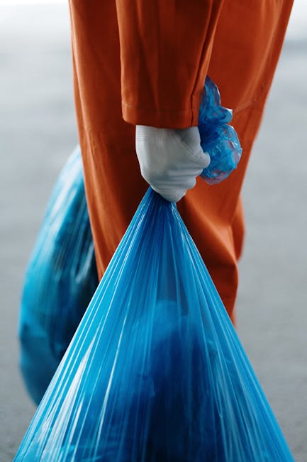 Close-up of a person wearing an orange-rust coloured protective jumpsuit and white gloves, holding a large, tightly knotted blue plastic rubbish bag by its tied top handle. The bag appears full and is made of durable, translucent plastic with a glossy finish, revealing some contents inside. The individual is standing outdoors on a pavement or concrete surface, with a blurred background suggesting an urban or industrial environment. The scene captures a moment of waste collection or rubbish removal, likely tied to private or alternative waste disposal services such as those offered by Rubbish Removal Hammersmith, with the focus on proper handling of waste materials in a professional manner.