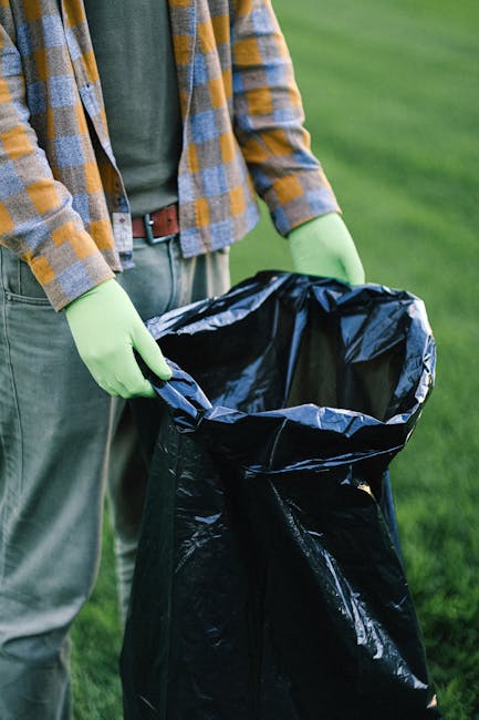 A person wearing a checkered yellow and blue shirt, with a dark green t-shirt underneath, is holding open a large black garbage bag lined with a shiny black plastic inner layer. The individual is wearing light green gloves for protection. They are standing outdoors on a grassy area with a well-maintained lawn visible in the background. The scene suggests they are participating in waste collection or rubbish removal, possibly as part of a private or independent waste disposal activity supported by companies like Rubbish Removal Hammersmith. The focus is on the bag and the person's hands, emphasizing the process of rubbish collection or on-site clearance, with natural lighting illuminating the scene, highlighting the textures of the materials and the vivid colors of the clothing and environment.