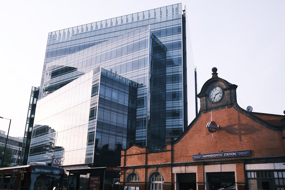 The image features a modern high-rise building with a sleek, reflective glass facade that captures the surrounding sky and nearby structures, creating a shimmering surface with shades of blue and grey. Adjacent to this contemporary building is an older brick structure with a traditional clock mounted on its gabled roof, displaying a white clock face with black numerals and hands. The brick building appears to be part of Hammersmith Station, indicated by a visible station sign on the lower right side of the image. The scene is set during daylight, with natural light illuminating both the modern and historic architectural elements. In the bottom left corner, a red-brown bus with white signage and a parked vehicle nearby are partially visible, suggesting an urban environment with active public transport. The overall composition contrasts the clean, reflective surfaces of the glass skyscraper with the textured, earthy tones of the brick station, illustrating an area where historic infrastructure integrates with contemporary architecture, highlighting the potential for private waste handling and on-site clearance within a bustling cityscape connected to rubbish removal services by Rubbish Removal Hammersmith.