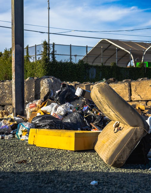 A close-up view of a pile of mixed waste and rubbish spilling onto a gravel surface in an outdoor environment. The collection includes black rubbish bags, some torn open revealing loose waste, a yellow plastic container, and an old, stained tyre leaning against the rubbish. Behind the refuse, there is a low stone wall made of irregularly shaped, rough-textured stones, partly obscured by the rubbish. In the background, a metal fence topped with wire mesh runs horizontally, with green foliage and trees visible beyond it. A tall metal pole with electrical wires extends vertically in the left portion of the image. To the right, part of an industrial structure with a curved roof and supporting framework can be seen, suggesting a commercial or storage site. The sky is mostly clear with some light clouds, giving the scene natural daylight. The overall setup indicates an area awaiting waste collection or an instance of improper waste disposal, relevant to private rubbish removal services such as those offered by Rubbish Removal Hammersmith.